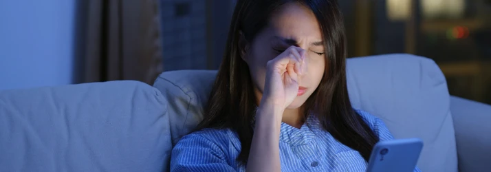 A young woman looking at her phone and rubbing at her irritated eye